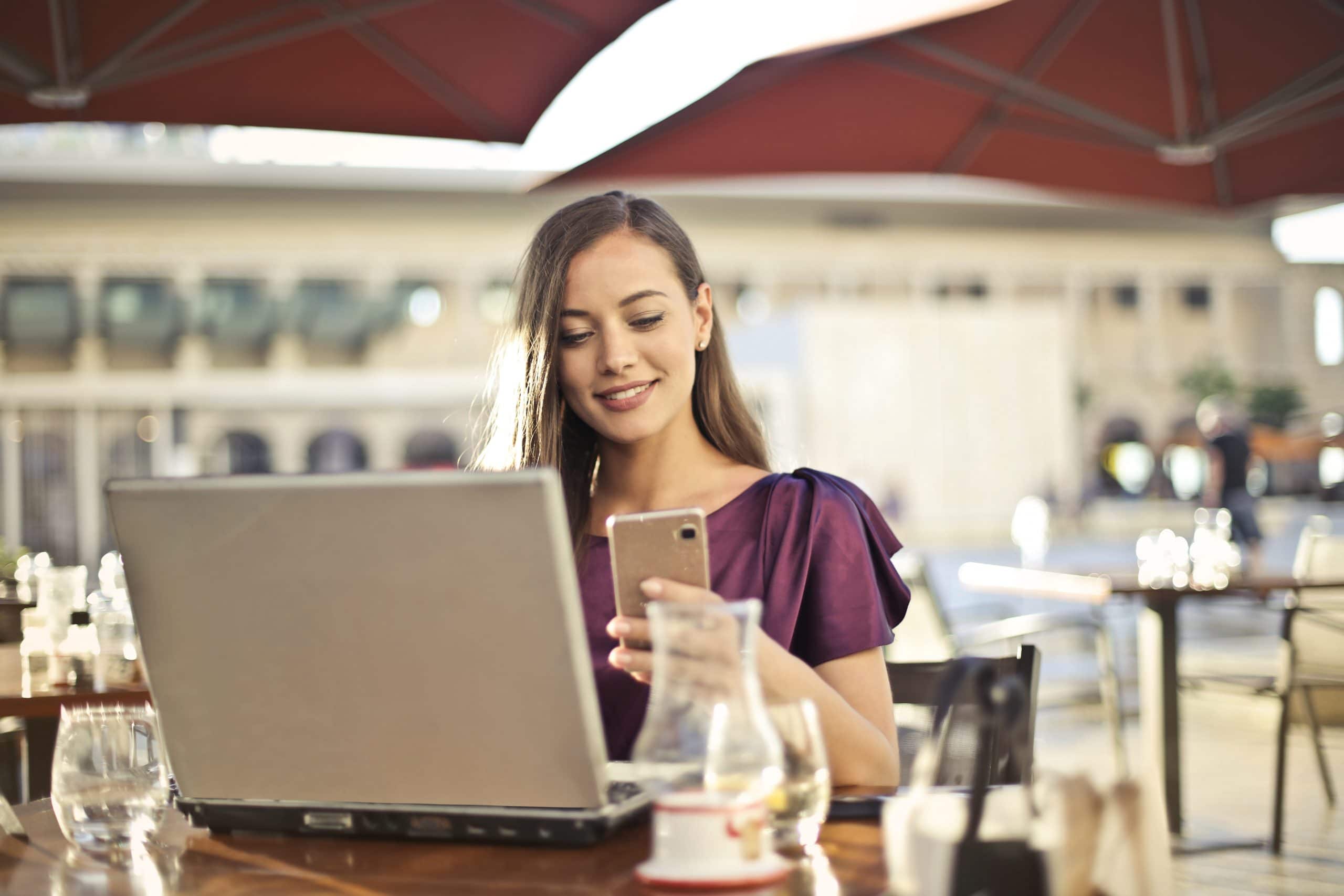 Mulher olhando para um notebook com celular na mão